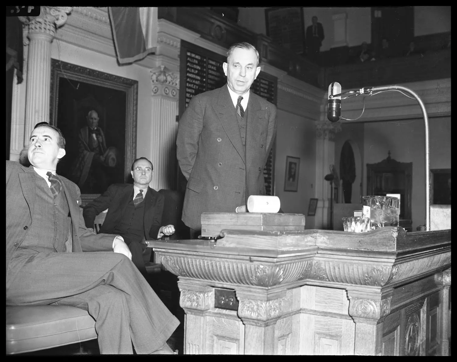 Black and white photo of a man in a suit speaking at a podium in a legislative chamber